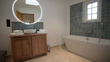 A modern bathroom with a wooden vanity, a round mirror with lights, a freestanding bathtub, and a window.