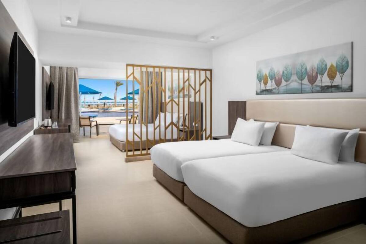 Ein Hotelzimmer mit Bett, Schreibtisch, Fernseher, Blick auf Pool und Strand sowie einem Bild an der Wand.