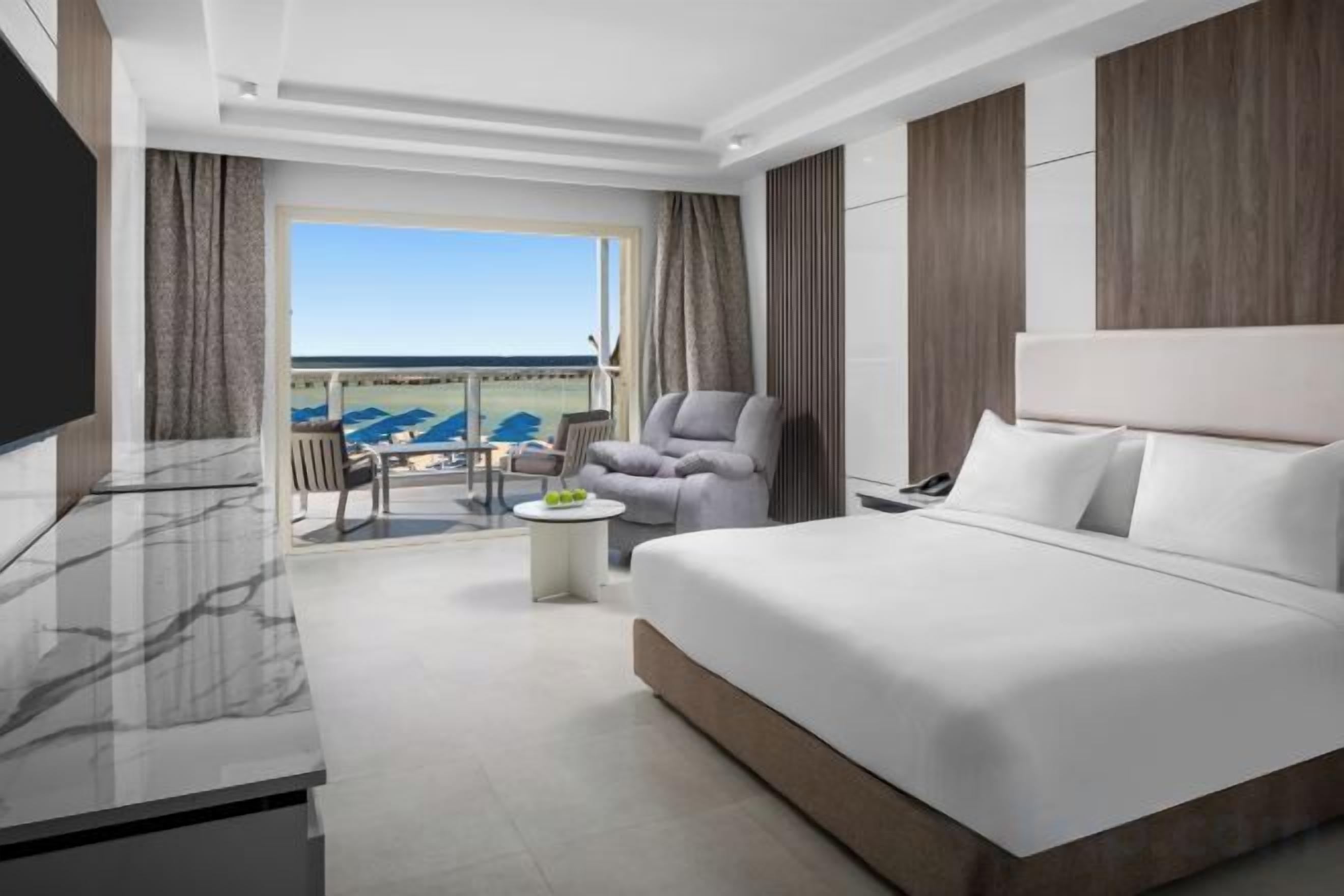 Habitación de hotel con una cama grande, televisor, balcón con vistas y un área de estar con mesa.