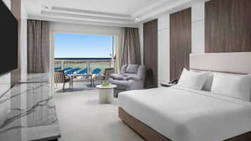 Habitación de hotel con una cama grande, televisor, balcón con vistas y un área de estar con mesa.