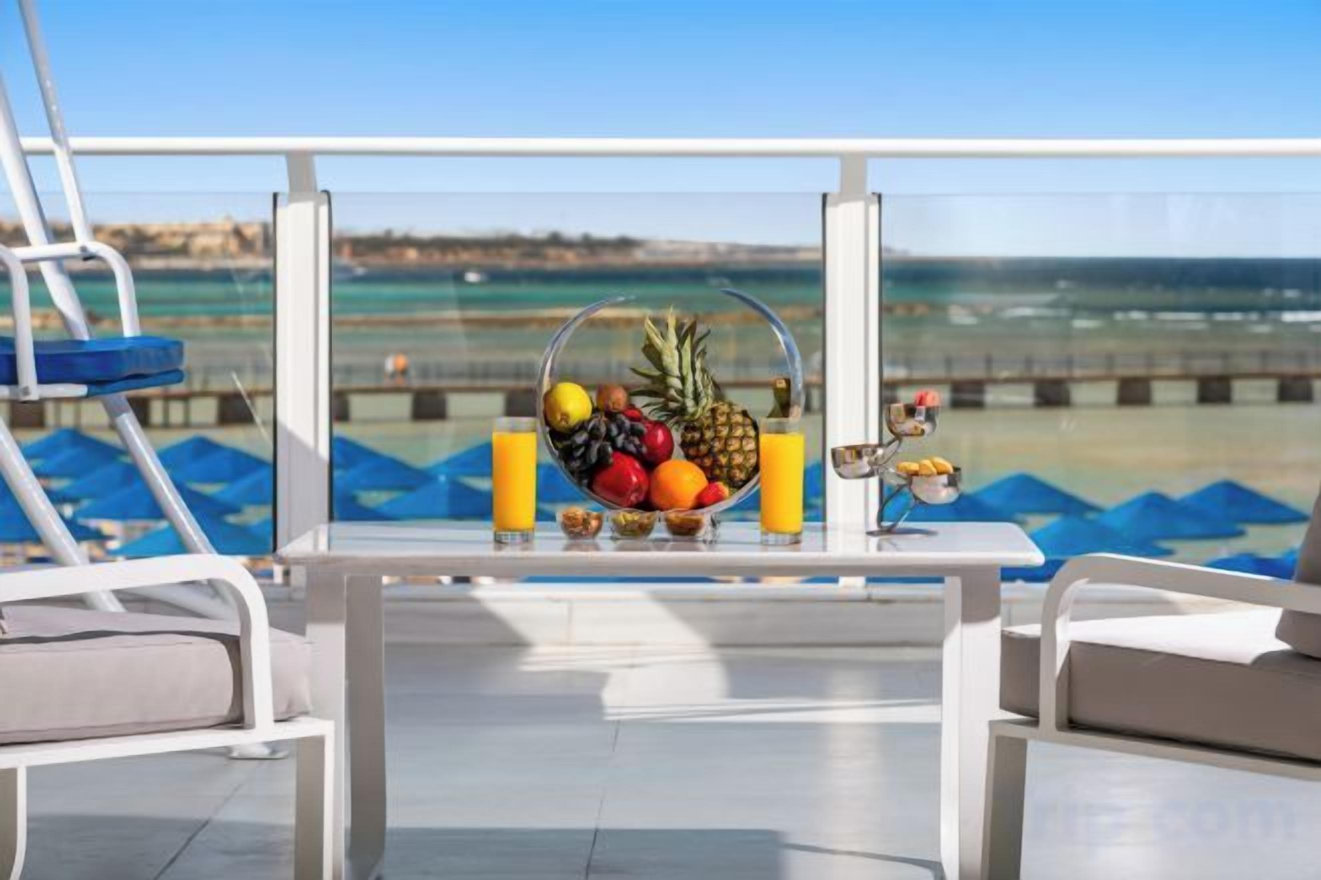A table with a fruit bowl, two glasses of orange juice, and a small bowl on a balcony overlooking the sea.