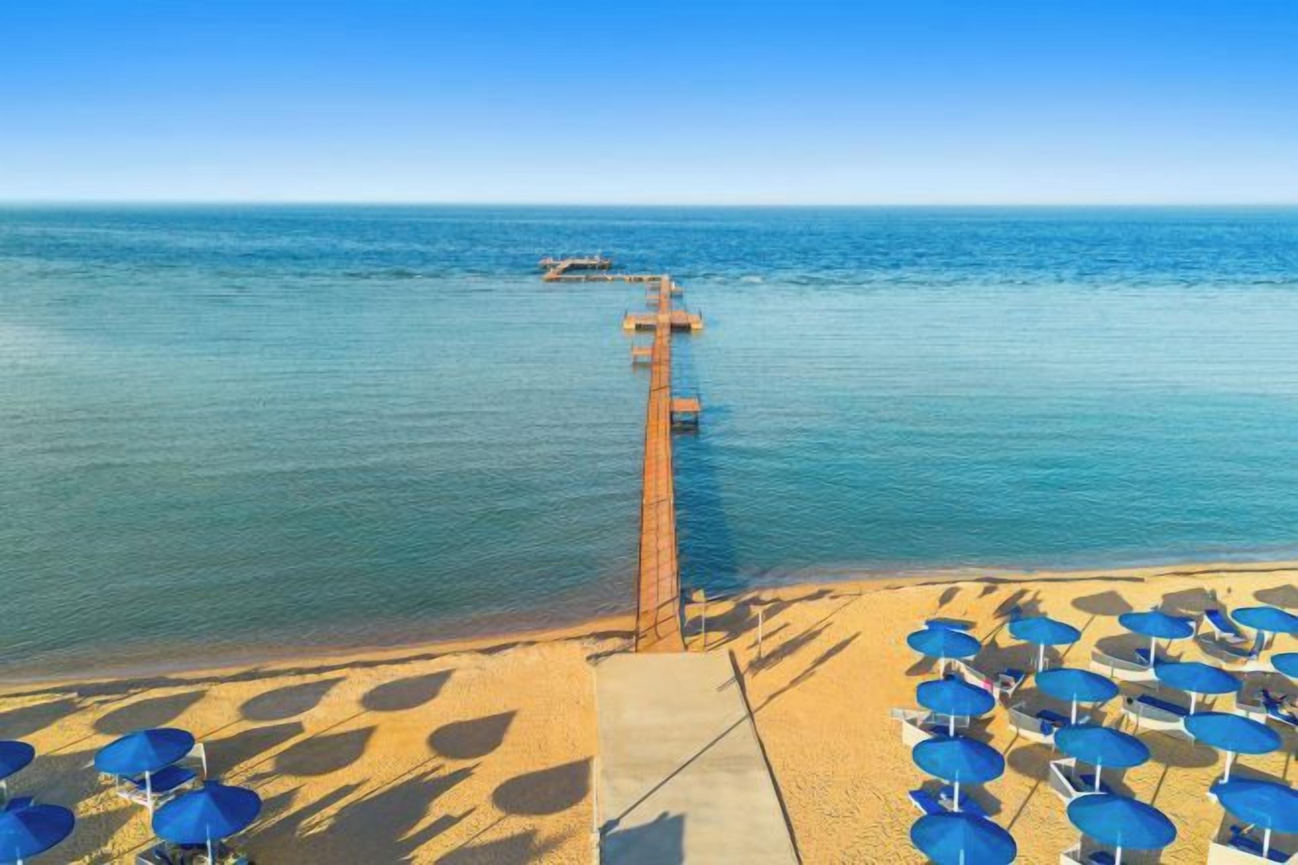 Een strand met een pier die het water in reikt, zandstrand met parasols en een heldere lucht.
