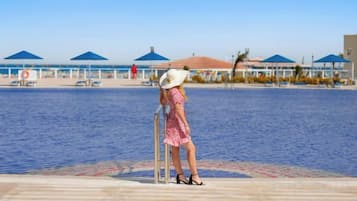 Mulher de chapéu branco e vestido rosa em deck de madeira à beira da água, com guarda-sóis azuis e espreguiçadeiras ao fundo.