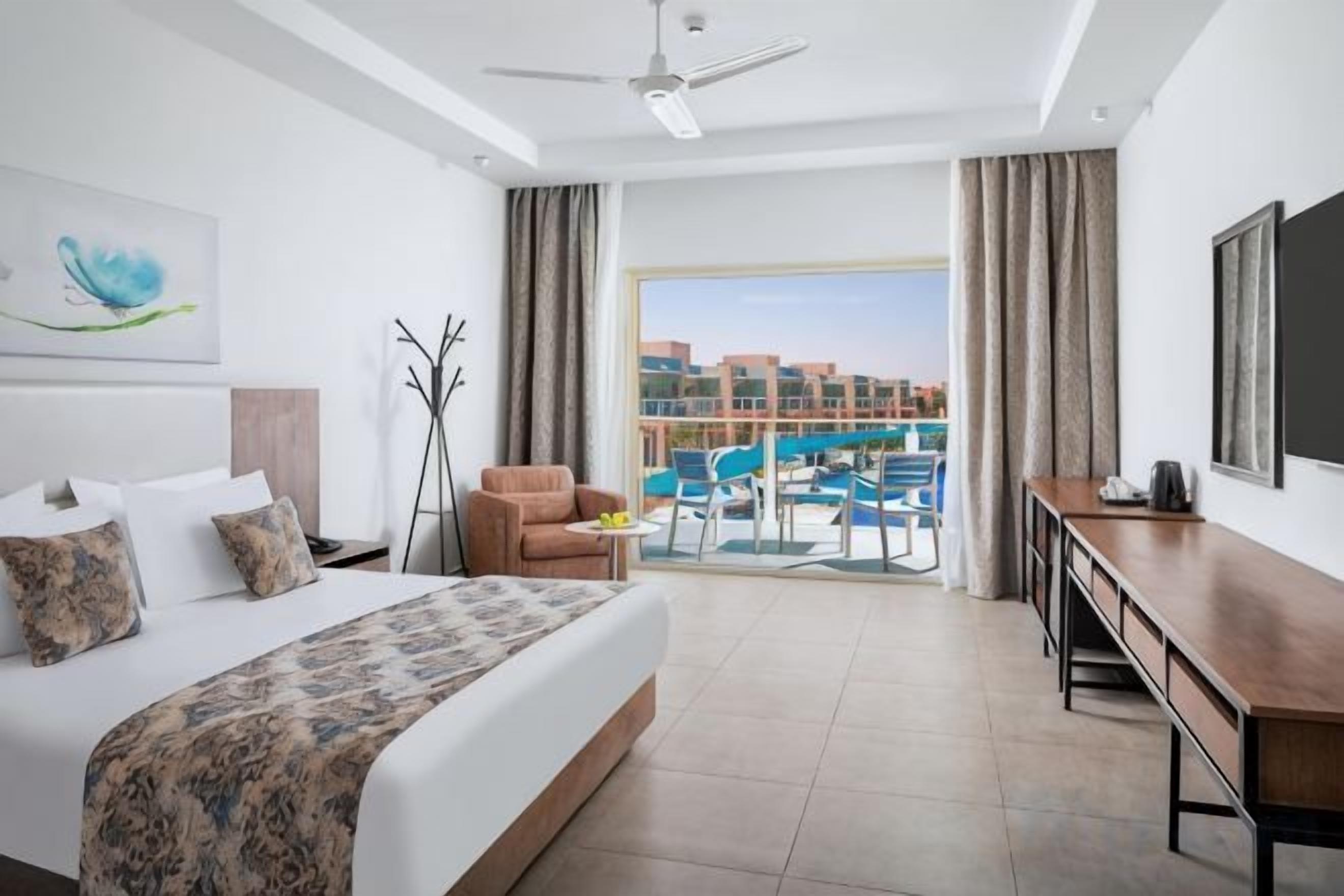 Quarto de hotel com cama, escrivaninha, cadeira, varanda com vista para a piscina e paisagem urbana ao fundo.