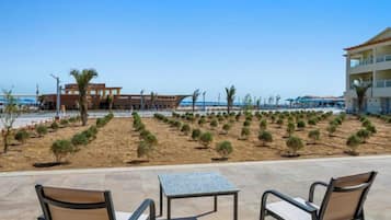 Een terras aan het strand met buitenmeubilair, een zandgebied met kleine planten, en een gebouw op de achtergrond.