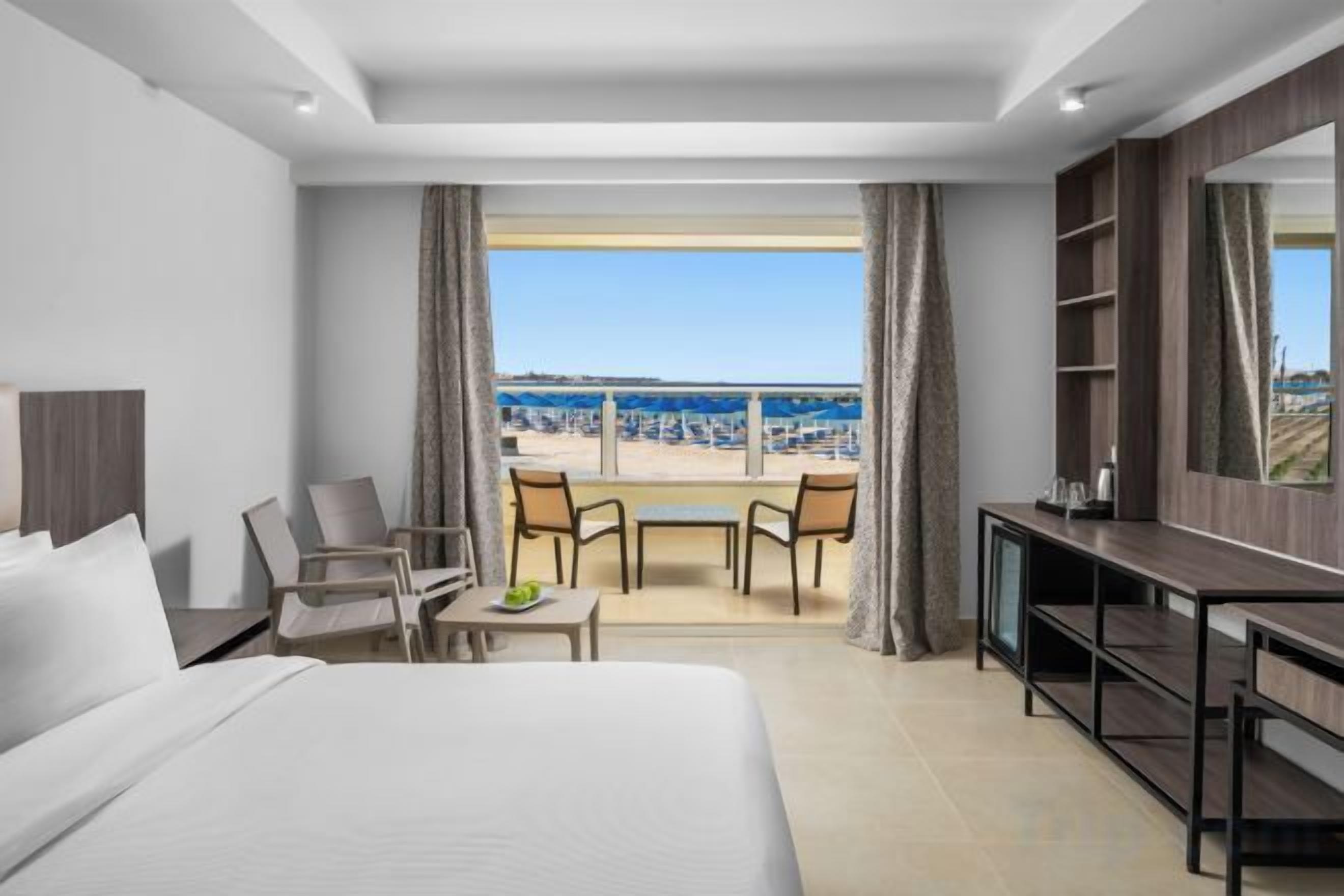 Ein Hotelzimmer mit einem großen Bett, einem Schreibtisch, einem Sessel, einem kleinen Tisch und einem Balkon mit Blick auf den Strand.