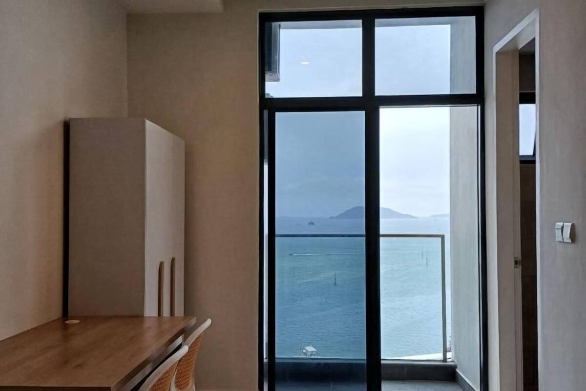 Une pièce comprenant une table en bois, un meuble et une grande porte-fenêtre coulissante donnant sur un balcon avec vue sur la mer et les montagnes.