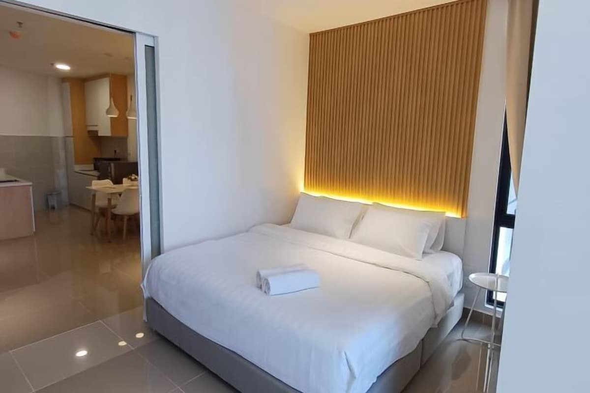 Una habitación de hotel moderna con una cama grande, una mesita de noche pequeña, una lámpara de pared y vistas al área de comedor contigua.