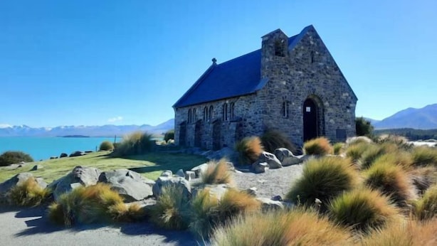 A stone church with a steep roof, surrounded by grassy hills and a clear blue sky. - Alpine Star (Lake Tekapo)