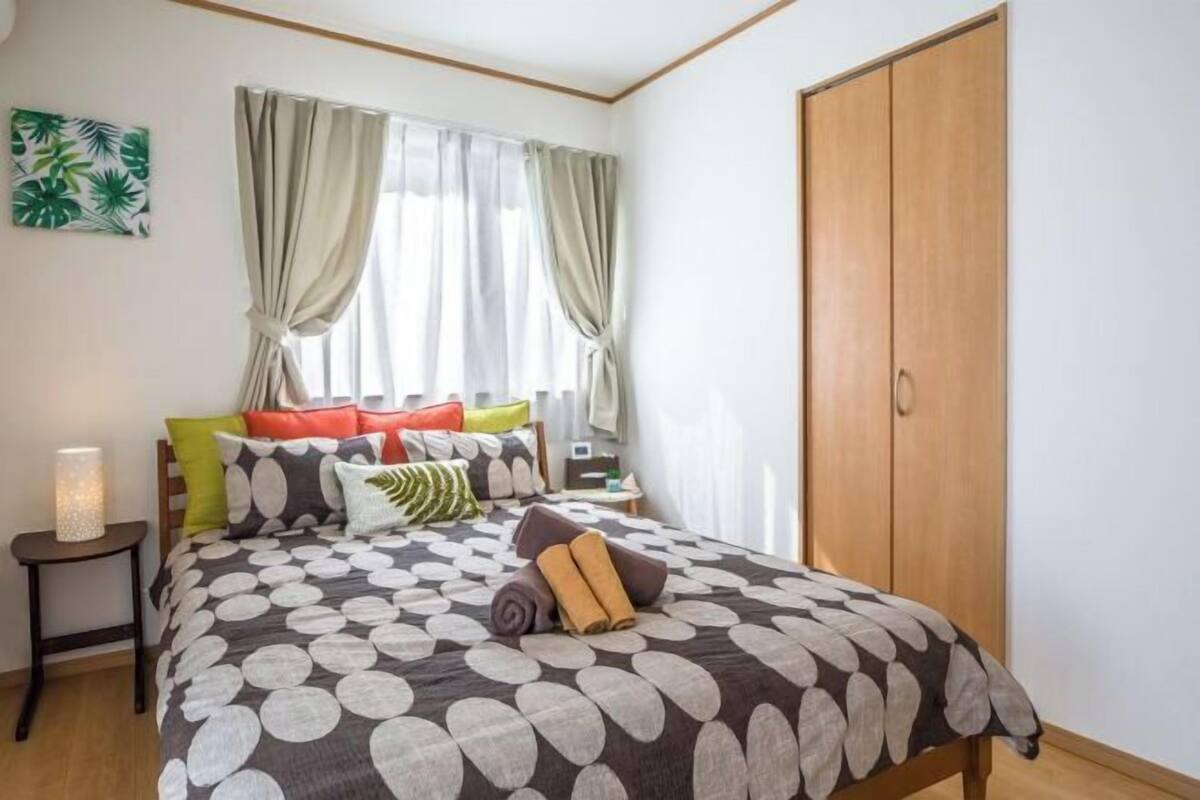 A bedroom with a bed, pillows, a nightstand, a lamp, a wardrobe, and a window with curtains.