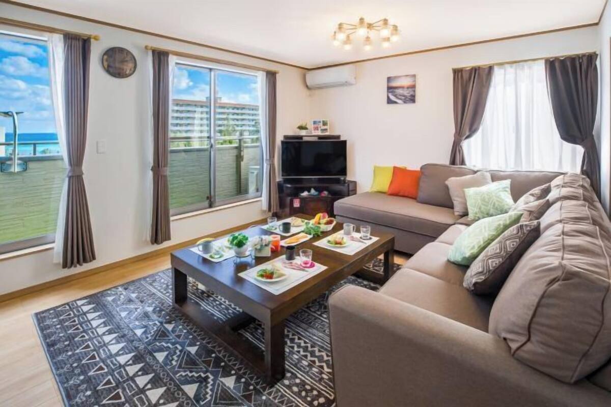 A living room with a sofa, a coffee table with food, a TV, and a balcony with curtains.