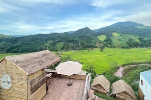 A thatched-roof building with a wooden deck and an umbrella, overlooking a lush green valley and mountains.