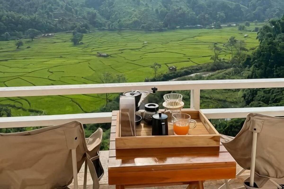 Una mesa de madera con una bandeja, una tetera, una taza y un vaso de zumo de naranja en un balcón con vistas a campos verdes.