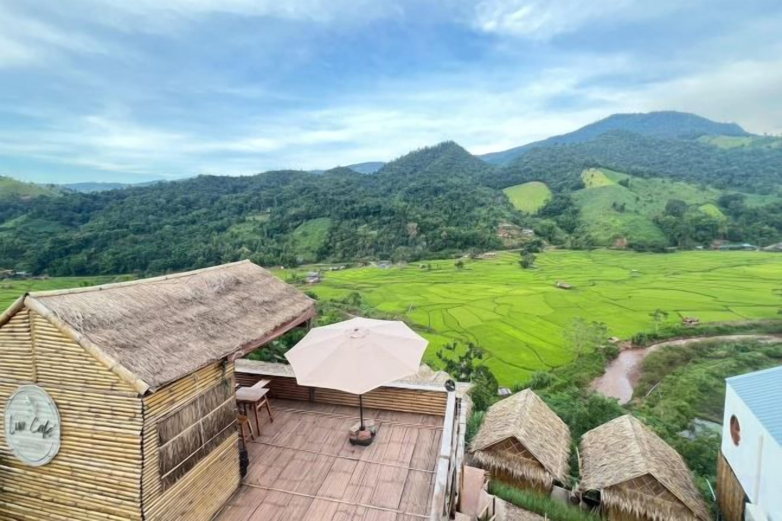 A wooden structure with a thatched roof, a table, and an umbrella, overlooking a green valley and hills.