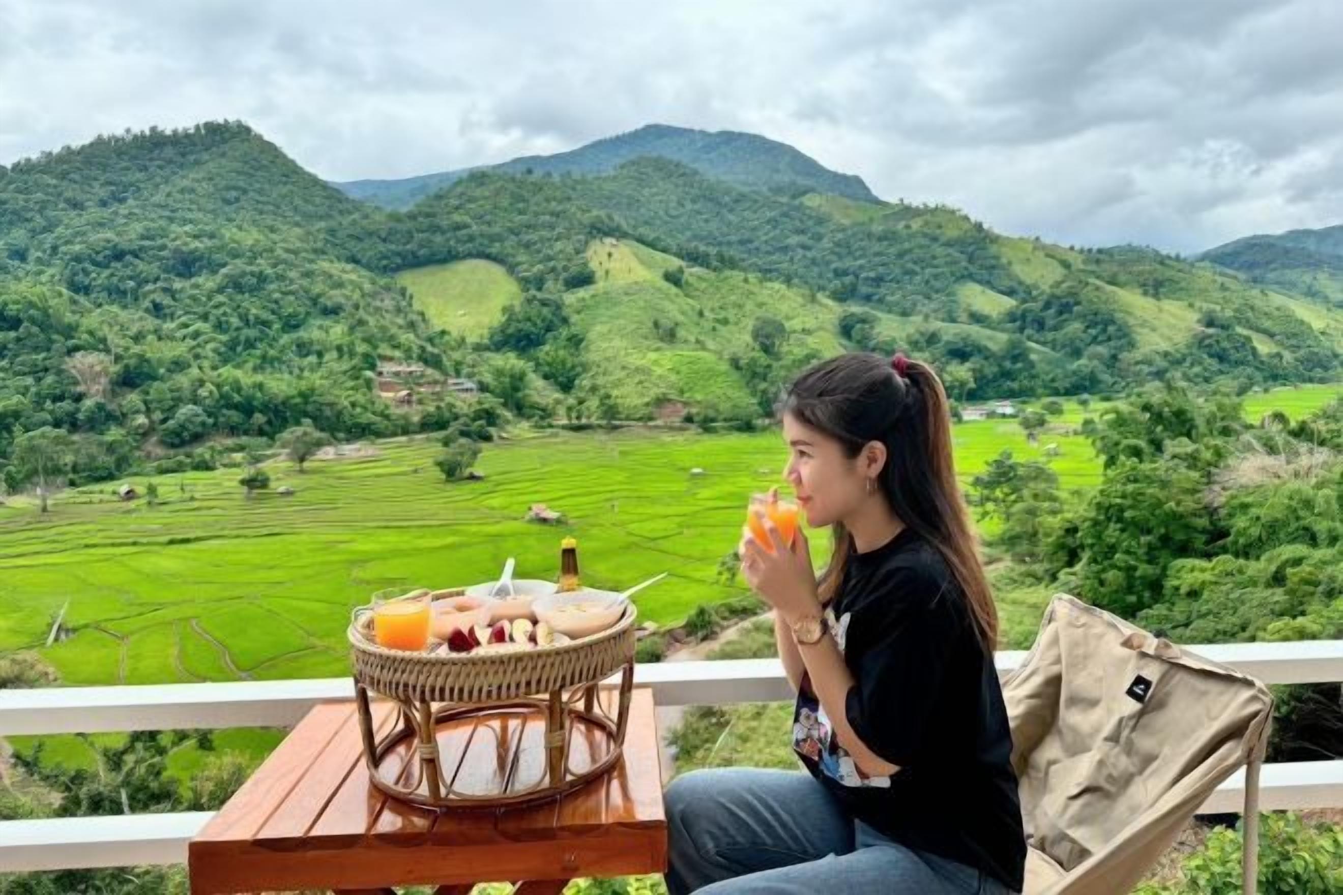 Una persona sentada en una mesa con una cesta de comida y una bebida, con vistas a un valle y colinas verdes.