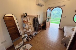 Una habitación con cama, suelo de madera, espejo, repisas con objetos y vista a través de una puerta abierta.