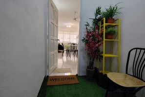 A hallway with a welcome mat, a chair, a yellow plant stand, and a dining area in the background. - Permai Ria Home Apartment (Tanjung Bungah)