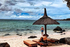 A beach with a thatched umbrella, lounge chairs, and a person walking towards the water. - Club Med Albion Villas (Mauritius)