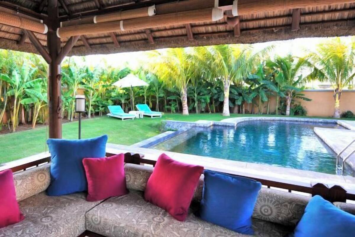 A pool area with a thatched roof, lounge chairs, and a seating area with cushions.