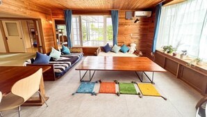 A cozy cabin-style room with a wooden ceiling, a large dining table, a sofa with cushions, and a window with curtains. - Base Shiraoi (Shiraoi)