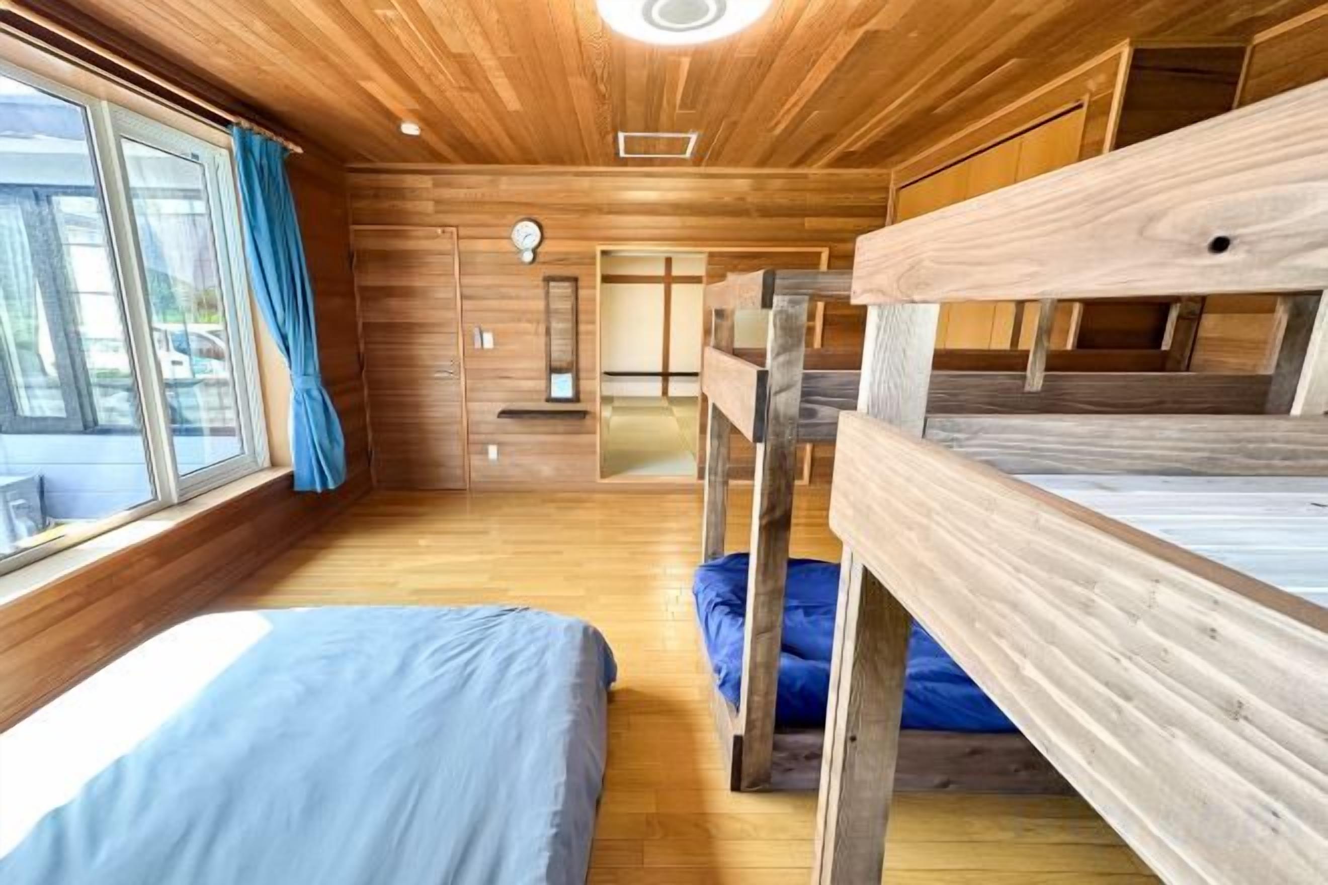 A room with bunk beds, a wooden floor, a window with curtains, and a door leading to another room.