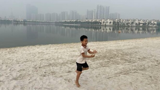 Un bambino gioca su una spiaggia sabbiosa con uno skyline urbano sullo sfondo.