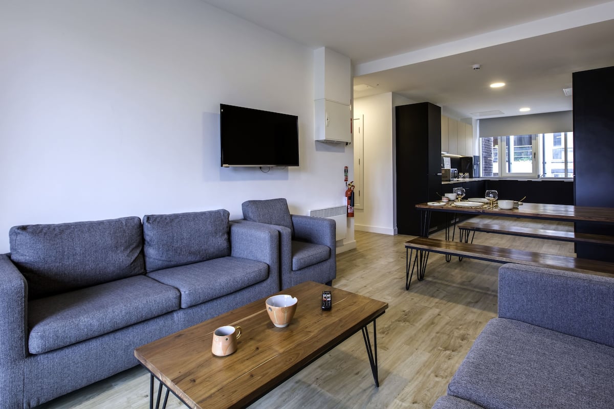 A modern living room with grey sofas, a wooden coffee table, and a flat-screen TV mounted on the wall.