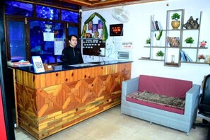 A reception desk with a wooden design, a calendar, and a digital clock. There is a sofa and a wall with shelves containing books and decorative items. - Dream Valley Kasol (Kasol)