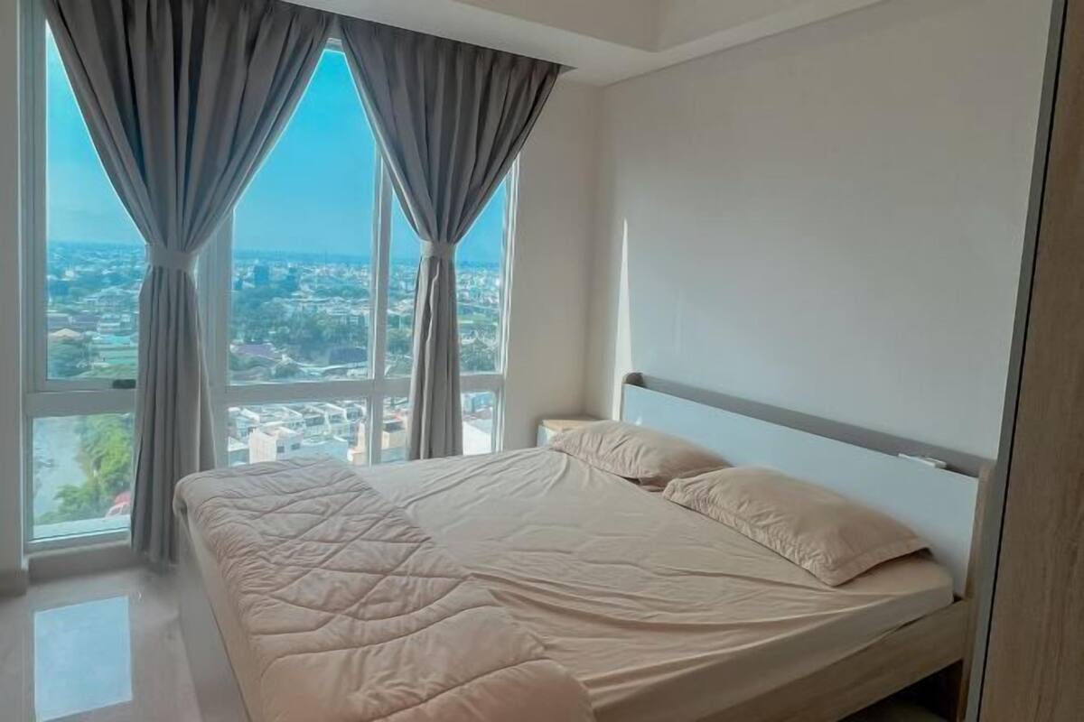 A bedroom with a bed, a view of a cityscape through large windows, and curtains.