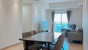 A modern living room with a large table and comfortable seating, a balcony with a city view, and a large window with curtains. - 3BR Podomoro Apartment @Empire Tower 107 sqm² (Kesawan)