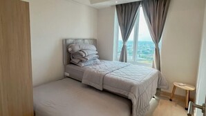 A modern bedroom with a large bed, a small stool, and a view of the city through the window. - 3BR Podomoro Apartment @Empire Tower 107 sqm² (Kesawan)