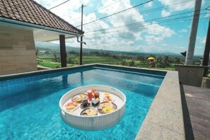 Una piscina con una zona de bar flotante, rodeada por un edificio con techo de tejas, y vistas a un paisaje verde.