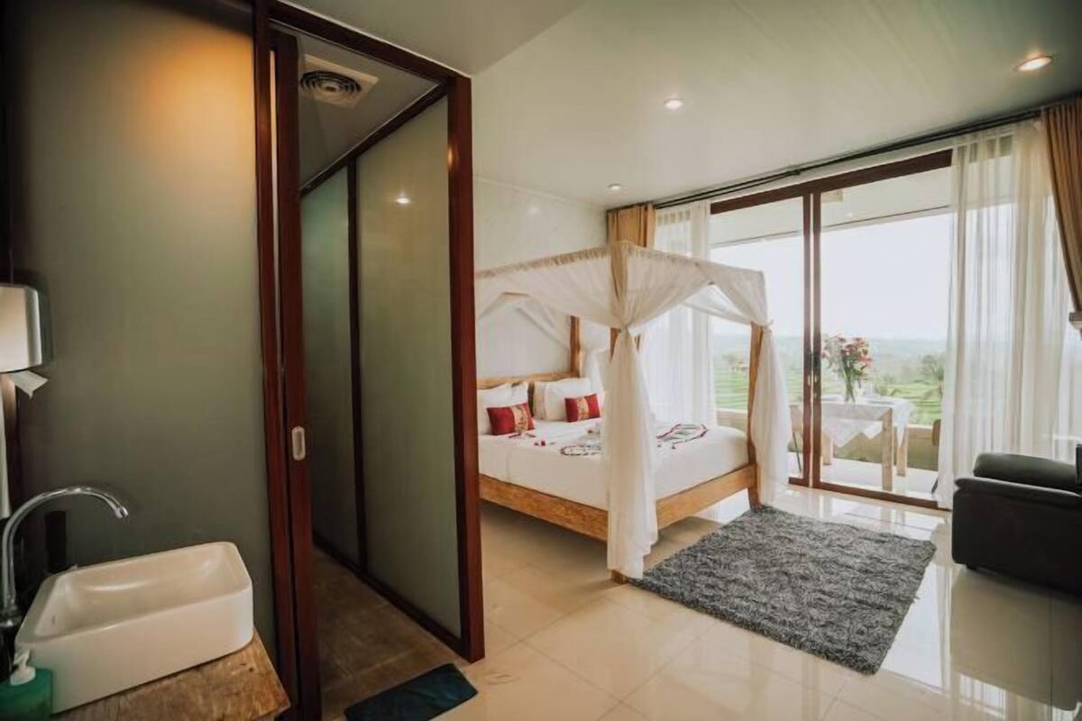 A bedroom with a bed, a large window, a balcony, and a bathroom.
