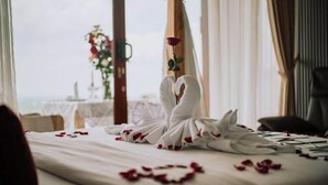 Habitación de hotel con una cama decorada con una elegante escultura de cisne hecha con una toalla blanca, p étalos de rosa esparcidos sobre la cama y vistas al mar a través de amplios ventanales.
