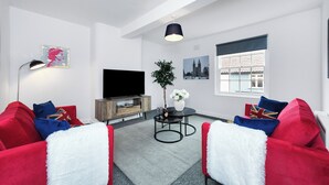 Una sala de estar moderna con un televisor de pantalla plana, sofás rojo y blanco, una pequeña mesa redonda con una planta y una ventana con persianas.