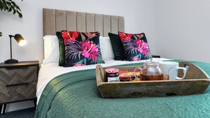 Una cama bien hecha con almohadillas estampadas, una lámpara de noche y una bandeja con tetera y tazas sobre una colcha verde.