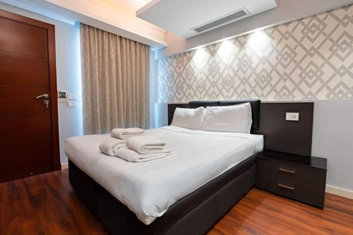 Une chambre d’hôtel avec un grand lit, une table de chevet sombre, une porte en bois et un mur à motifs.