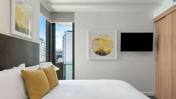 A modern hotel room with a large bed, a flat-screen TV mounted on the wall, and a city view through the window. - Quattro on Astor (Spring Hill)