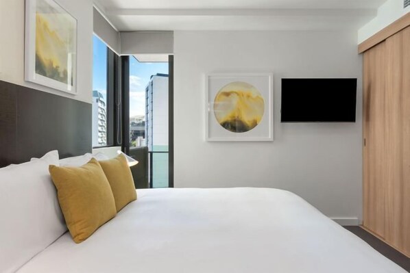 A modern hotel room with a large bed, a flat-screen TV mounted on the wall, and a city view through the window.
