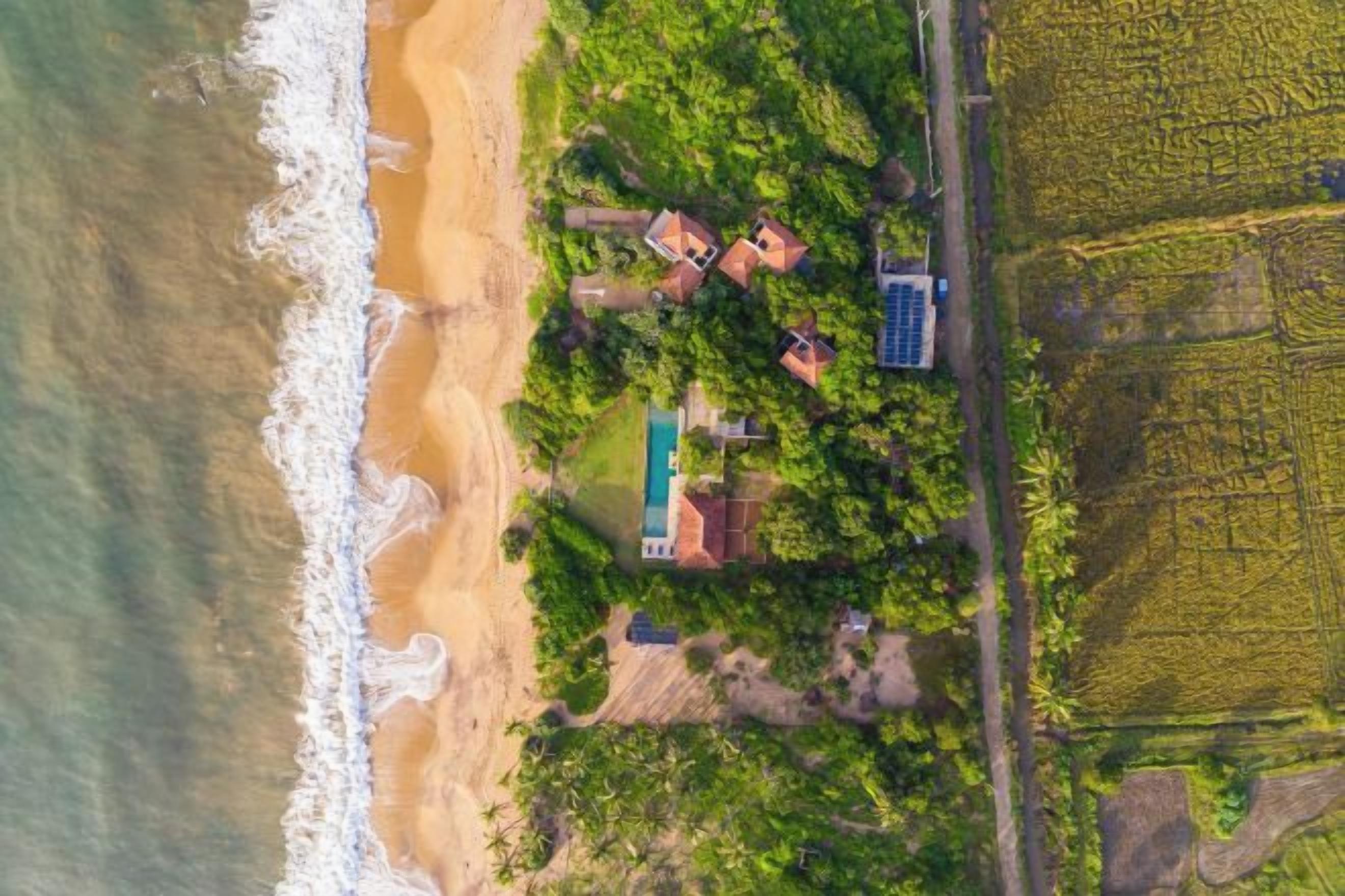 An aerial view of a coastal property with a beach, a swimming pool, and surrounding vegetation.