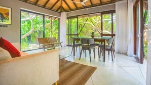 A spacious room with a high ceiling, large windows offering a view of lush greenery, a dining area with a table and chairs, and a ceiling fan. - Ravana Garden (Hambantota)