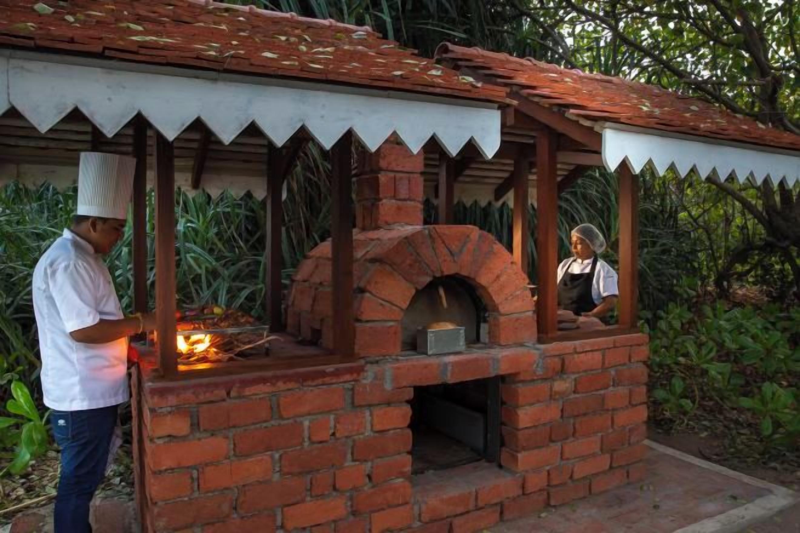 A brick-built outdoor oven with a chimney, a person cooking inside, and another person observing.
