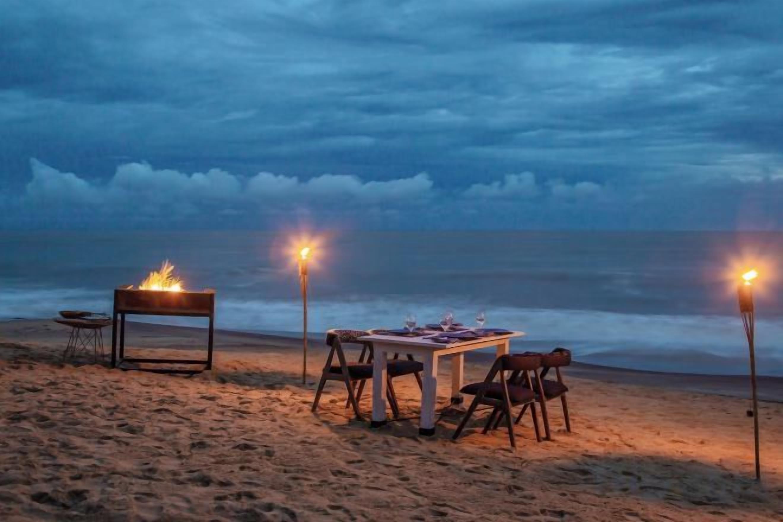 A beachside dining setup with a table and chairs, a fire pit, and two torches providing light.