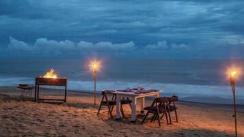 A beachside dining setup with a table and chairs, a fire pit, and two torches providing light.