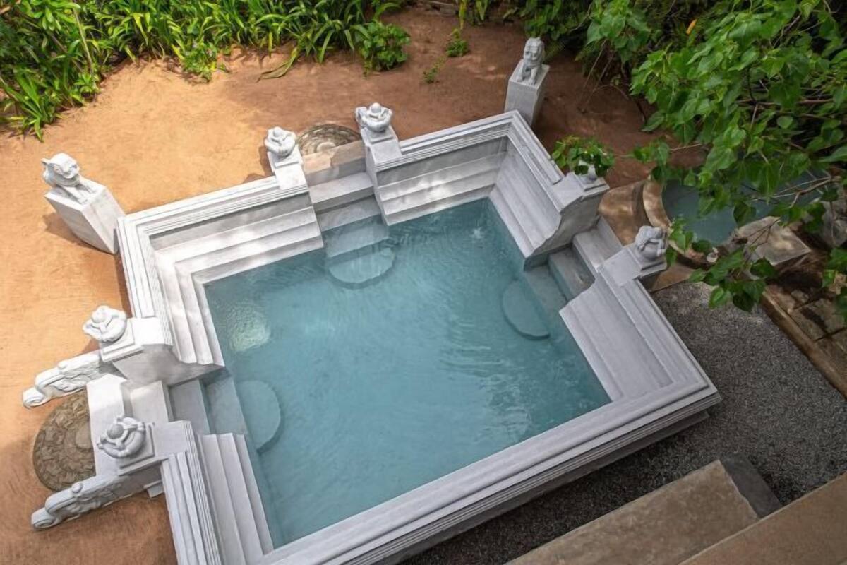 A square-shaped pool with stone statues at each corner, surrounded by a sandy area and greenery.