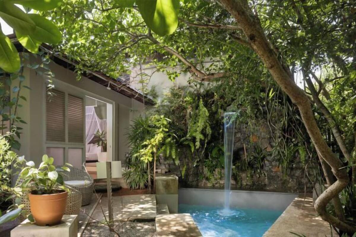A private pool with a waterfall, surrounded by lush greenery and a house with a balcony.