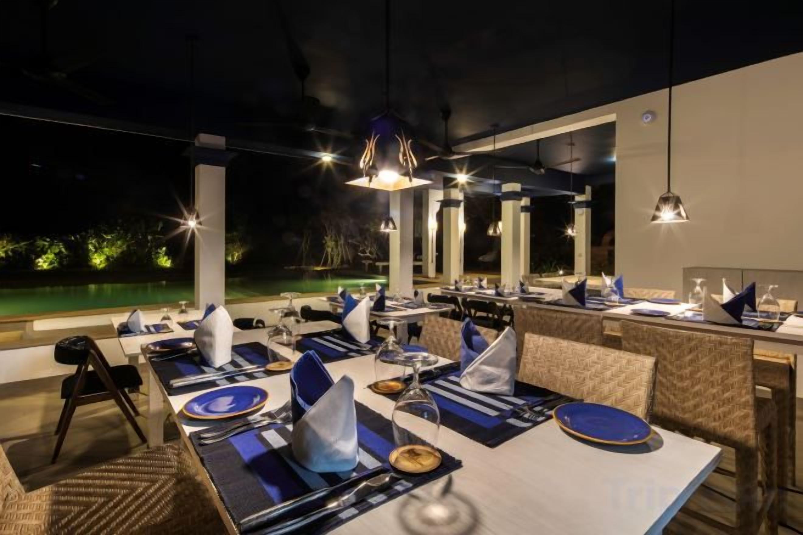 An outdoor dining area with tables set for two, overhead lighting, and a view of a pool.