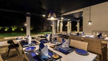 An outdoor dining area with tables set for two, overhead lighting, and a view of a pool.