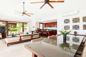 Una sala de estar moderna con una mesa de comedor de vidrio, un sofá, ventiladores de techo y una cocina con armarios y encimera.
