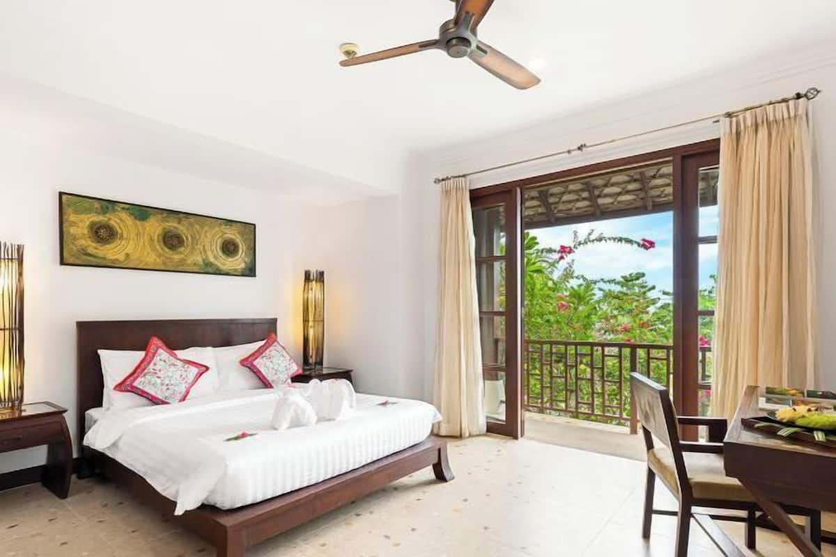 Camera d'albergo con un letto, una ventola a soffitto, un balcone affacciato sul verde e un tavolo con una sedia.